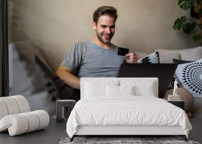 Happy young man using laptop sitting on sofa shopping online at home in the living room Wall mural